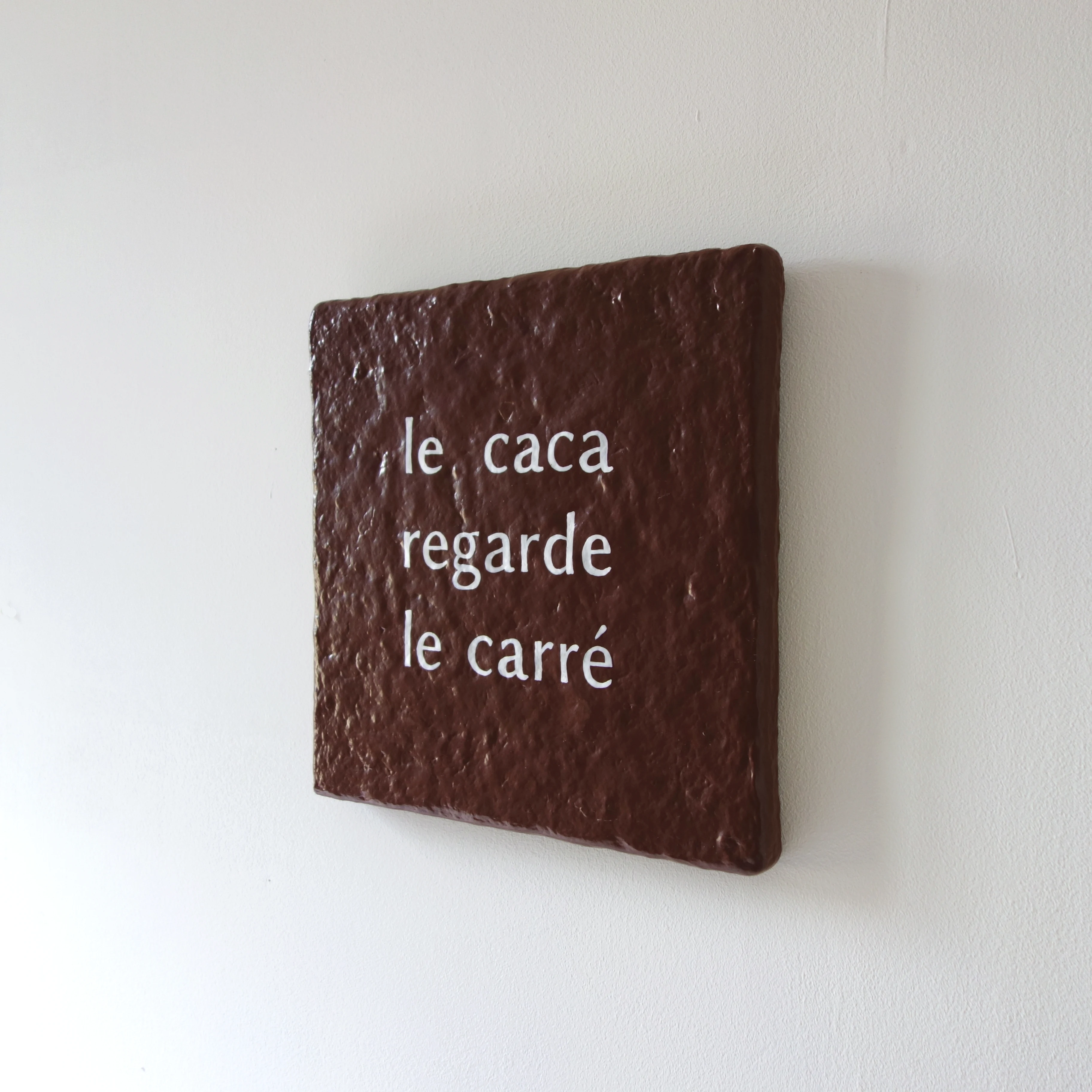 Un tableau carré en verre peint en rouge rosé sur l'envers. La peinture est grattée pour écrire : le caca regarde le carré. Vue de face.