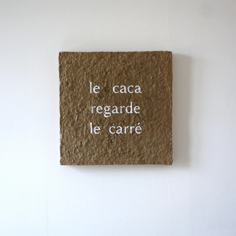 Un tableau carré en verre peint en bleu sur l'envers. La peinture est grattée pour écrire : le caca regarde le carré. Vue de face