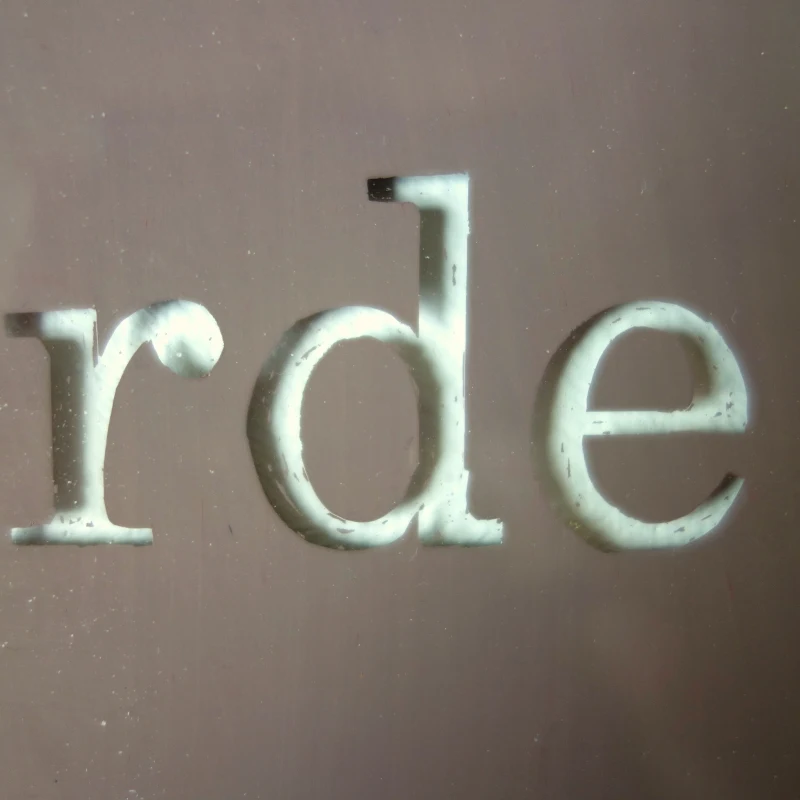 Un tableau carré en verre peint en brun sur l'envers. La peinture est grattée pour écrire : le caca regarde le carré. Vue de détail.