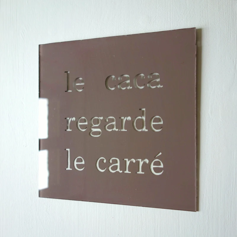 Un tableau carré en verre peint en brun sur l'envers. La peinture est grattée pour écrire : le caca regarde le carré. Vue de trois-quarts.