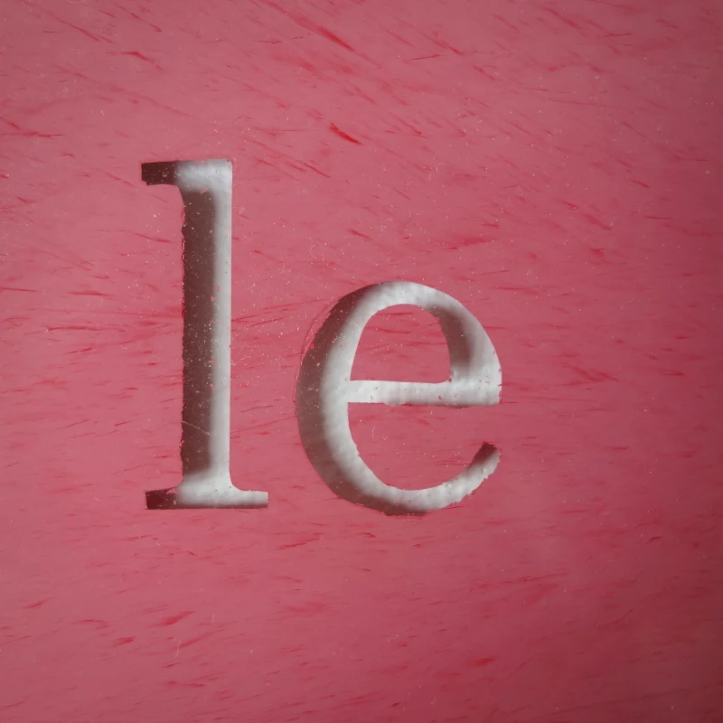 Un tableau carré en verre peint en rouge rosé sur l'envers. La peinture est grattée pour écrire : le caca regarde le carré. Vue de détail.