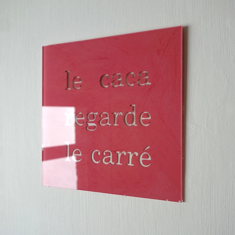 Un tableau carré en verre peint en rouge rosé sur l'envers. La peinture est grattée pour écrire : le caca regarde le carré. Vue de trois-quarts.